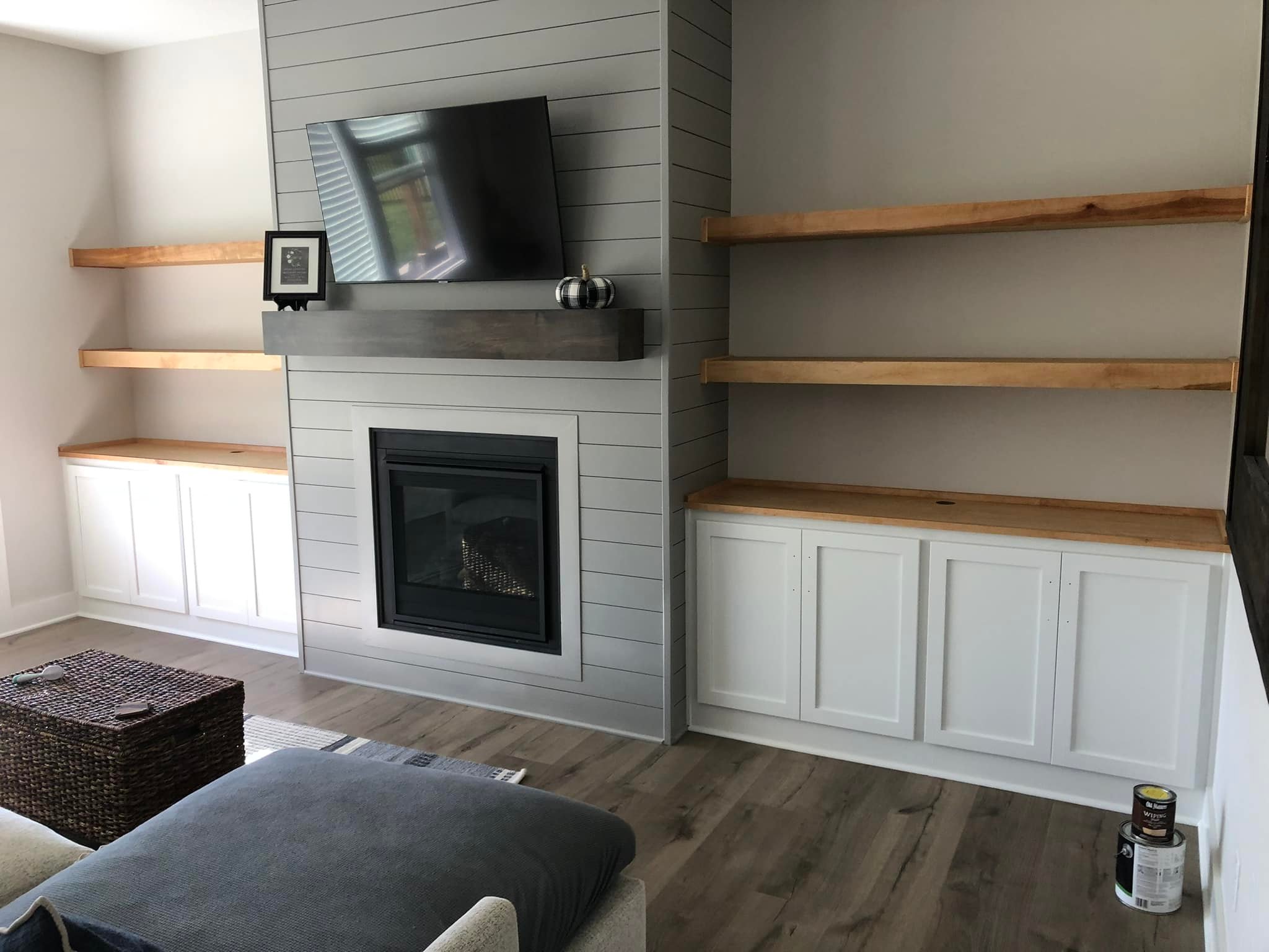 Living room with a sleek gray shiplap wall, a mounted TV, and a black fireplace. Wooden shelves and white cabinets flank each side. Cozy atmosphere.