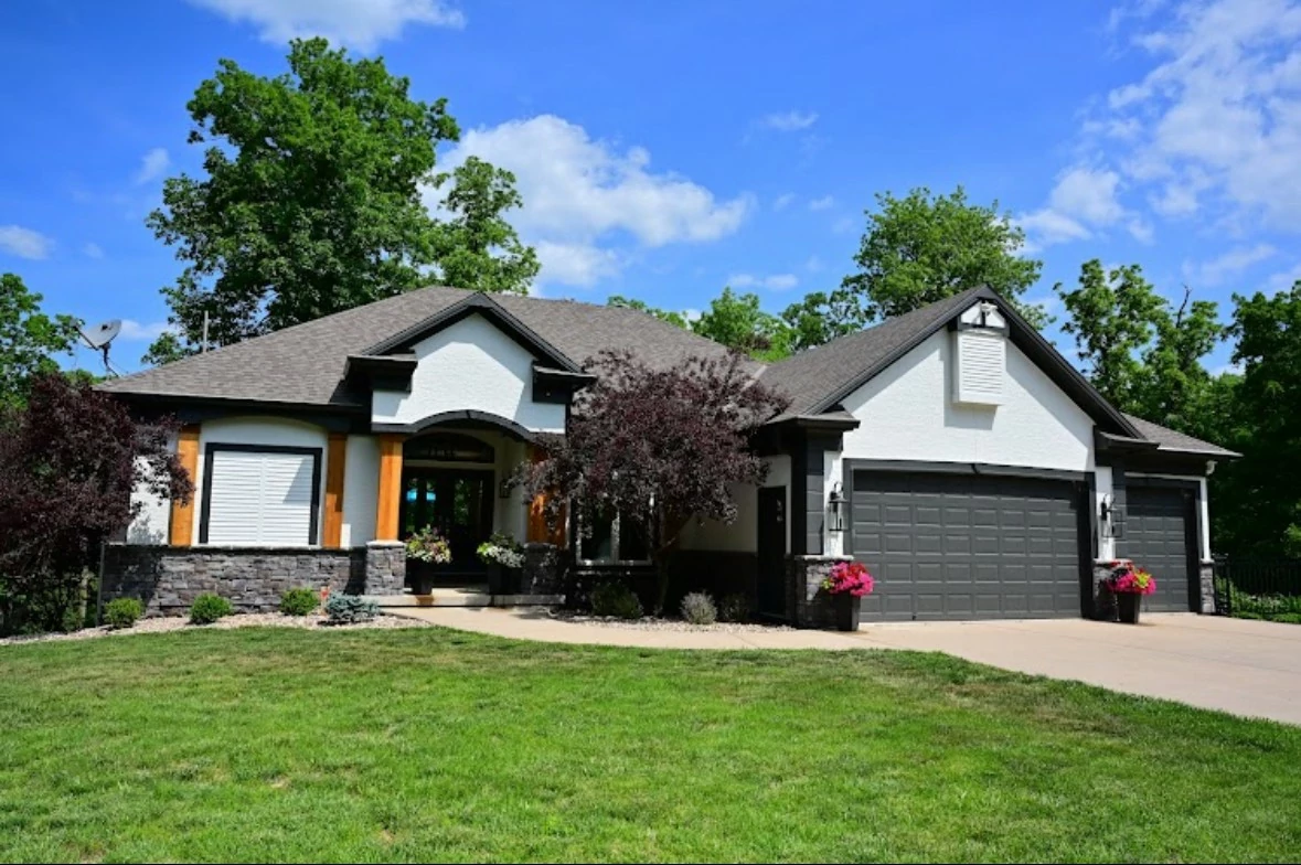 A modern suburban house with a gray roof and white exterior, surrounded by lush trees and a manicured lawn. The driveway leads to a three-car garage.